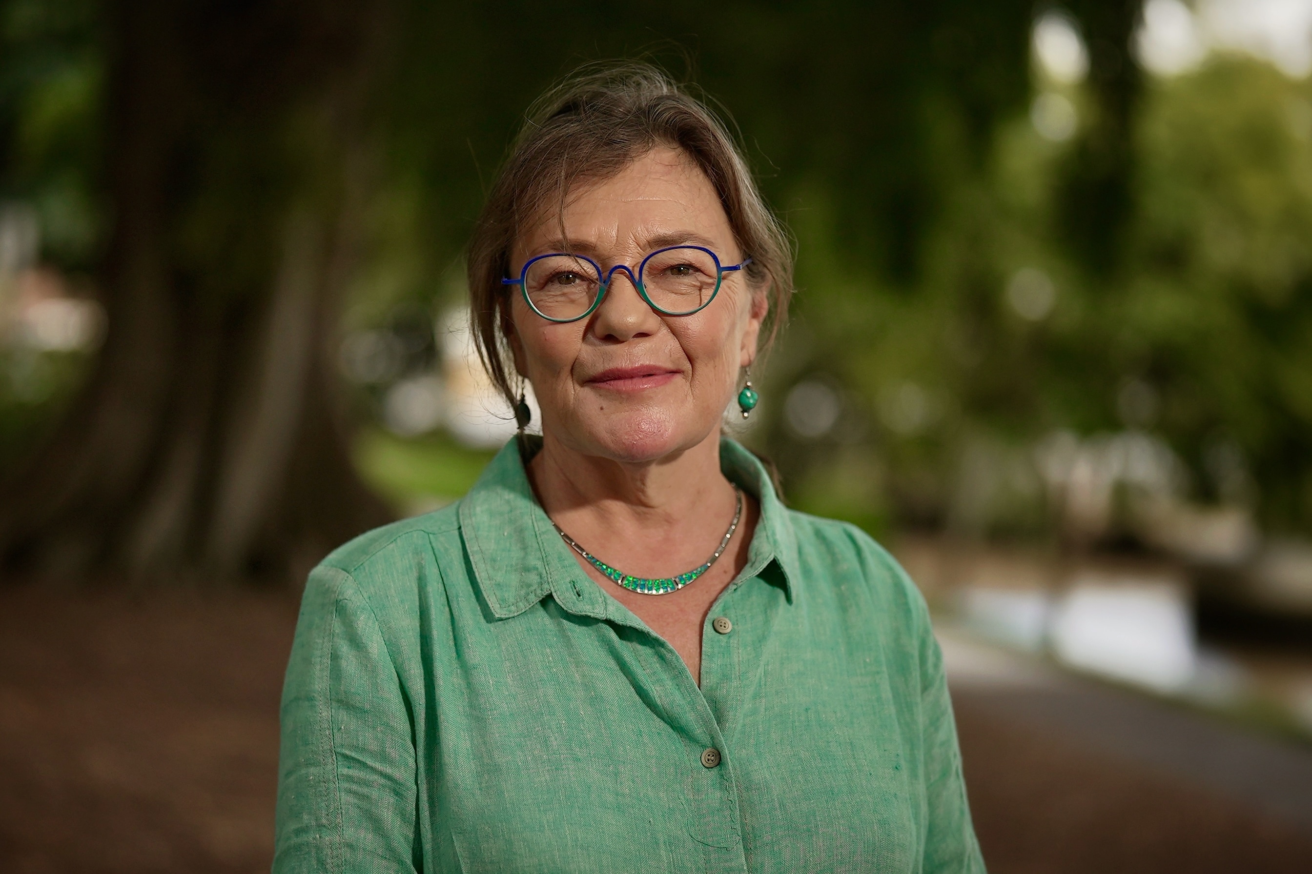 Mujer de mediana edad con camisa verde, collar y gafas mirando a la cámara, río borroso y árbol detrás de ella