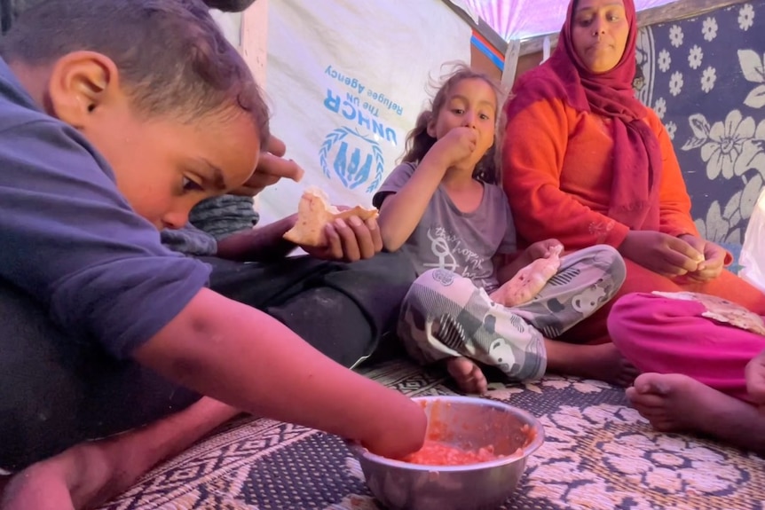 Several young children and a woman sitting on the ground inside a tent sharing one bowl of food.