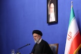 A man with a white beard, wearing a black turban, sits at a desk in front of a framed portrate of Ayatollah Khomeini.