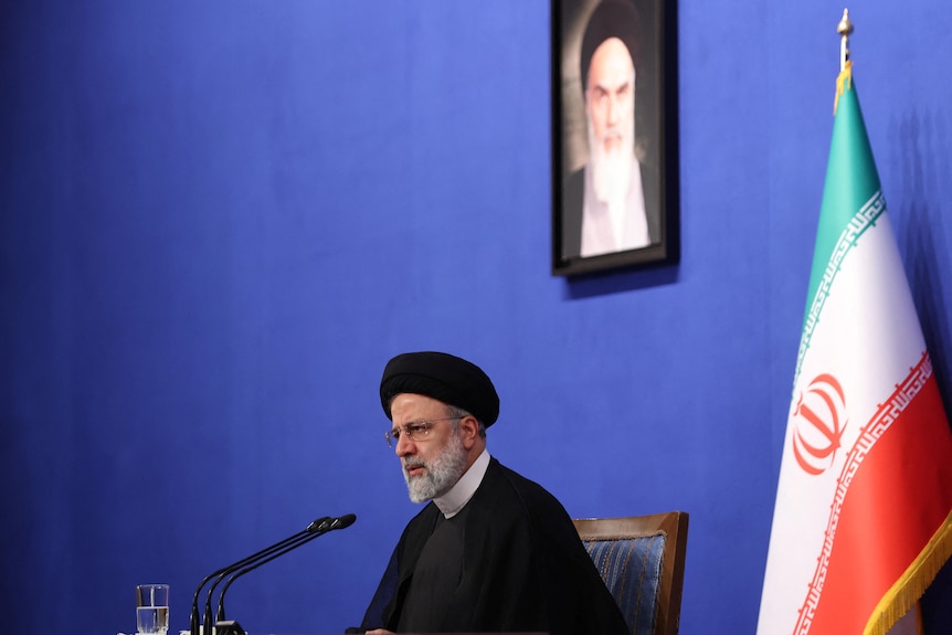 A man with a white beard, wearing a black turban, sits at a desk in front of a framed portrate of Ayatollah Khomeini.