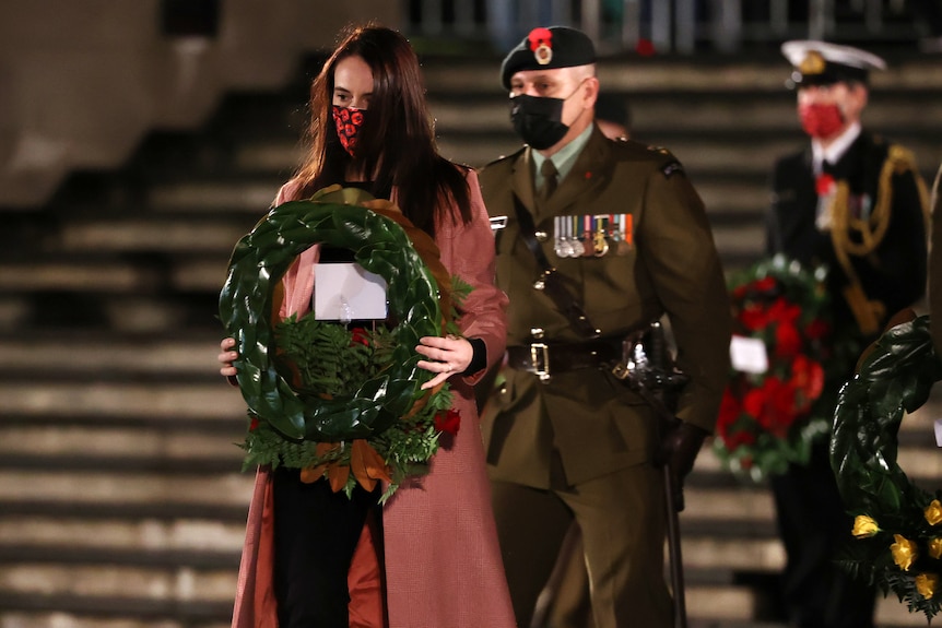 new zealand pm jacinda ardern holds a green commemorative wreath while wearing a face mask with red poppies on it
