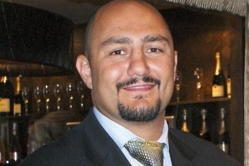 a bald man in a suit smiling in front of a wine cellar