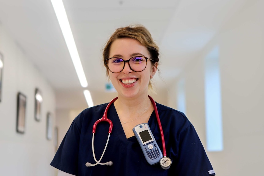 Nurse Anna Kidman wearing glasses and navy blue scrubs and stethoscope standing in illuminated hospital corridor