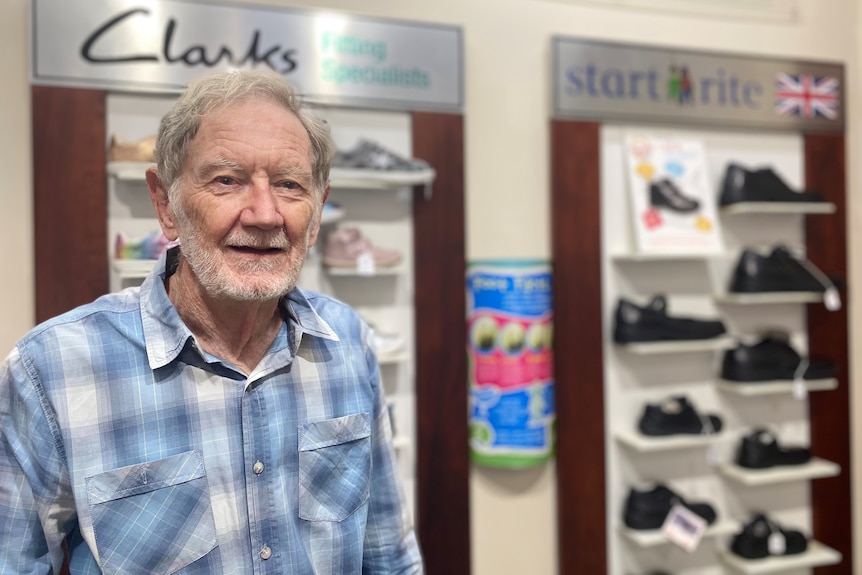 A man with white hair and a white beard, wearing a check-shirt, stands in front of a shoe display.