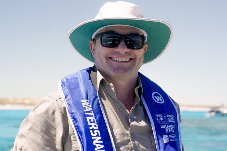 Miles Parsons, with wide-brimmed white hat, smiles widely, surrounded by ocean, holding a large, tube-shaped object.