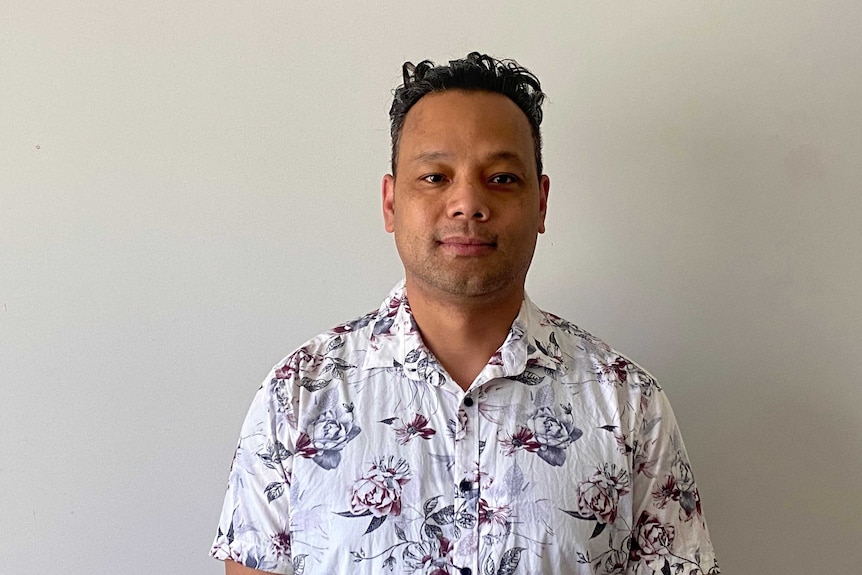 A Solomon Islander man wearing a white floral shirt stands in front of a white wall