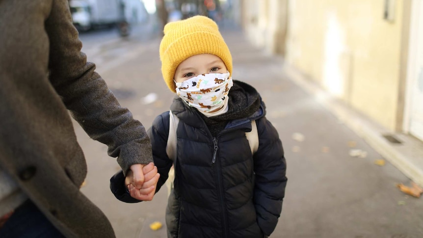 A young girl in a yellow beanie and an animal facemask standing on the street holding an unseen adult's hand
