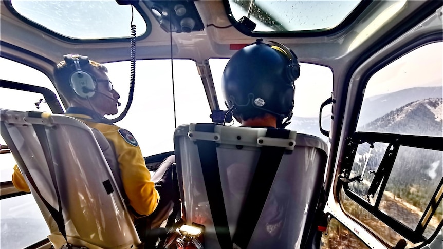 James Koens sits alongside another pilot in a helicopter flying over a bushfire.