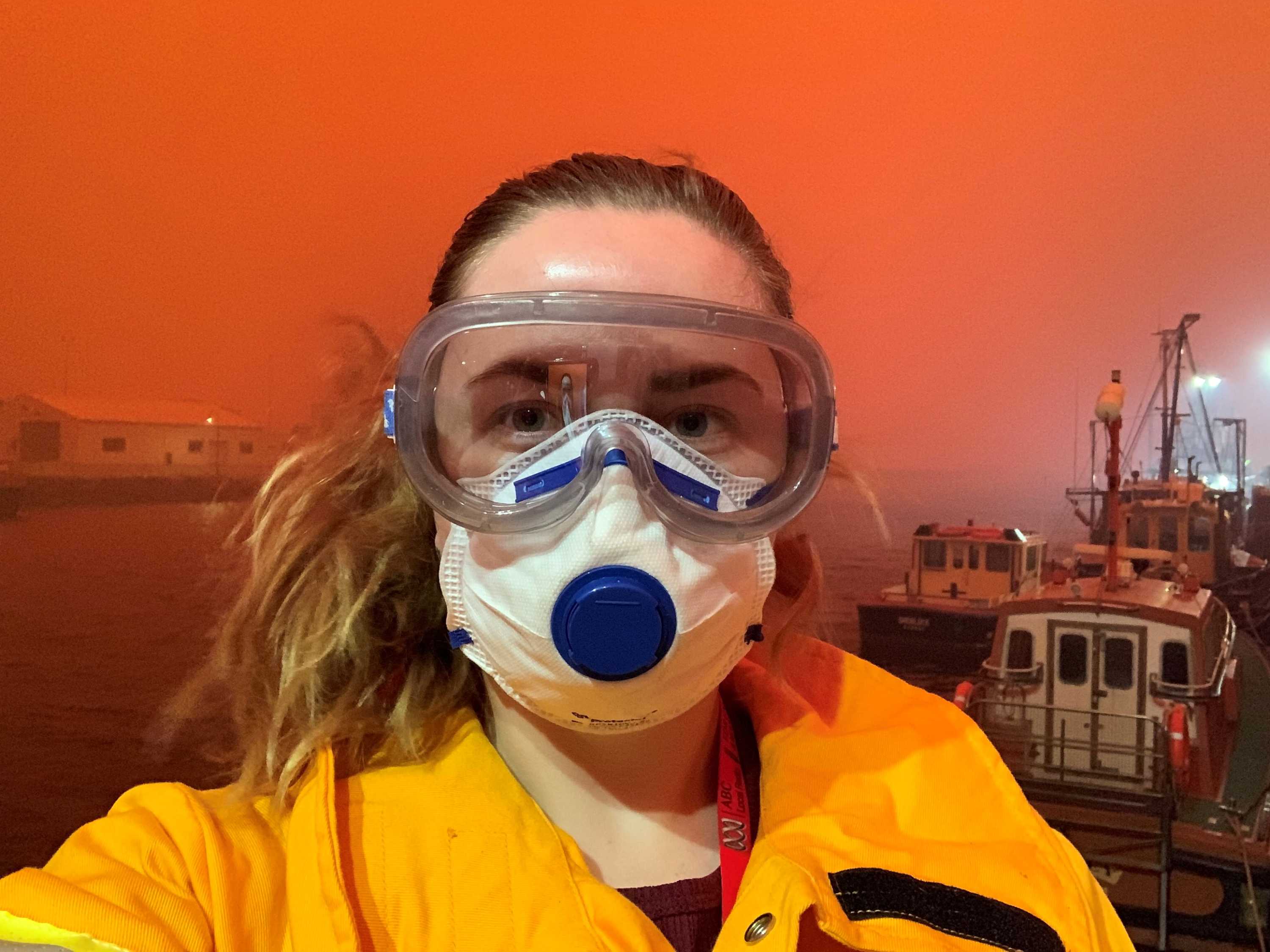 Head shot of Reardon wearing goggles and face mask with wharf and bright orange sky in background.