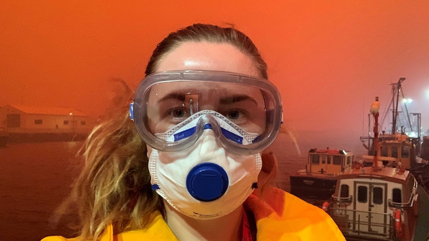 Head shot of Reardon wearing goggles and face mask with wharf and bright orange sky in background.