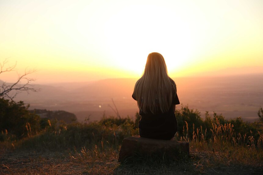 A woman with her back to the camera looks at a sunset from high on a hill.