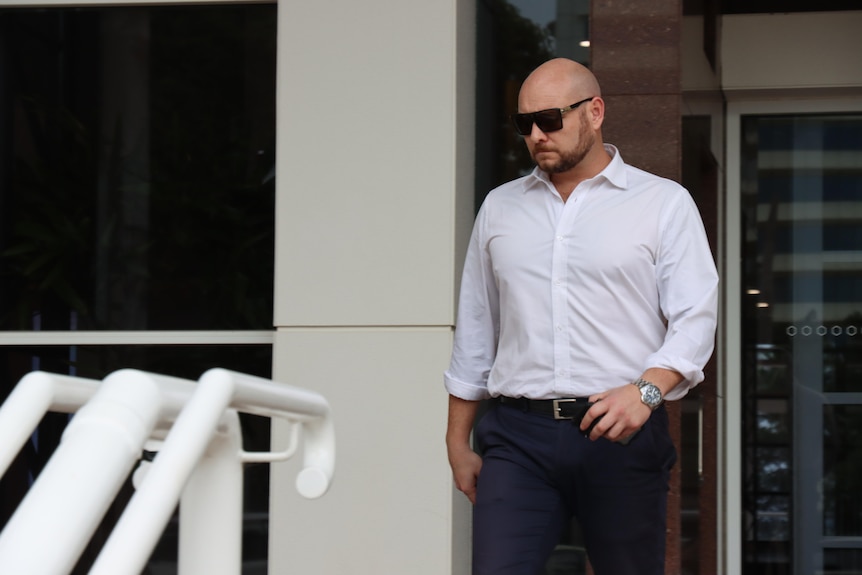 A man wearing a white business shirt and sunglasses walks alone from a courthouse building. 
