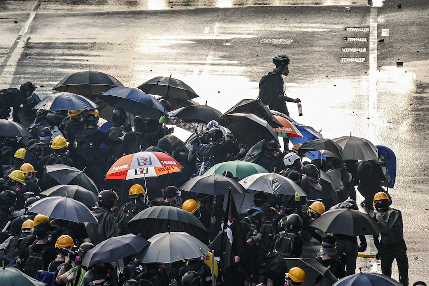 A crowd of protesters in helmets and wielding umbrella on a street