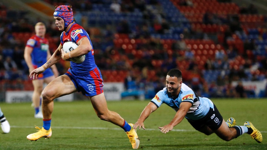 A Newcastle Knights NRL player runs with the ball tucked under his left arm as a Cronulla opponent dives to tackle him.