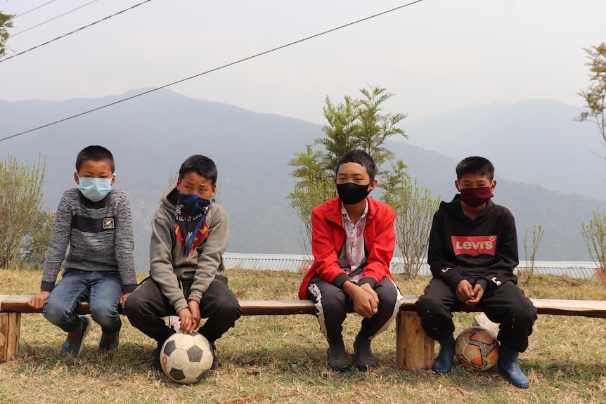 A row of young boys in face masks sit on a bench with mountains behind them