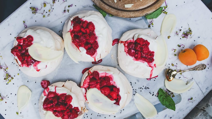 Five mini pavlovas with cream, white chocolate and raspberries presented on a marble table.