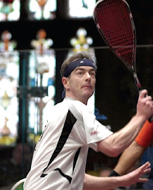 A middle aged man in white prepares to play squash backhand on a court