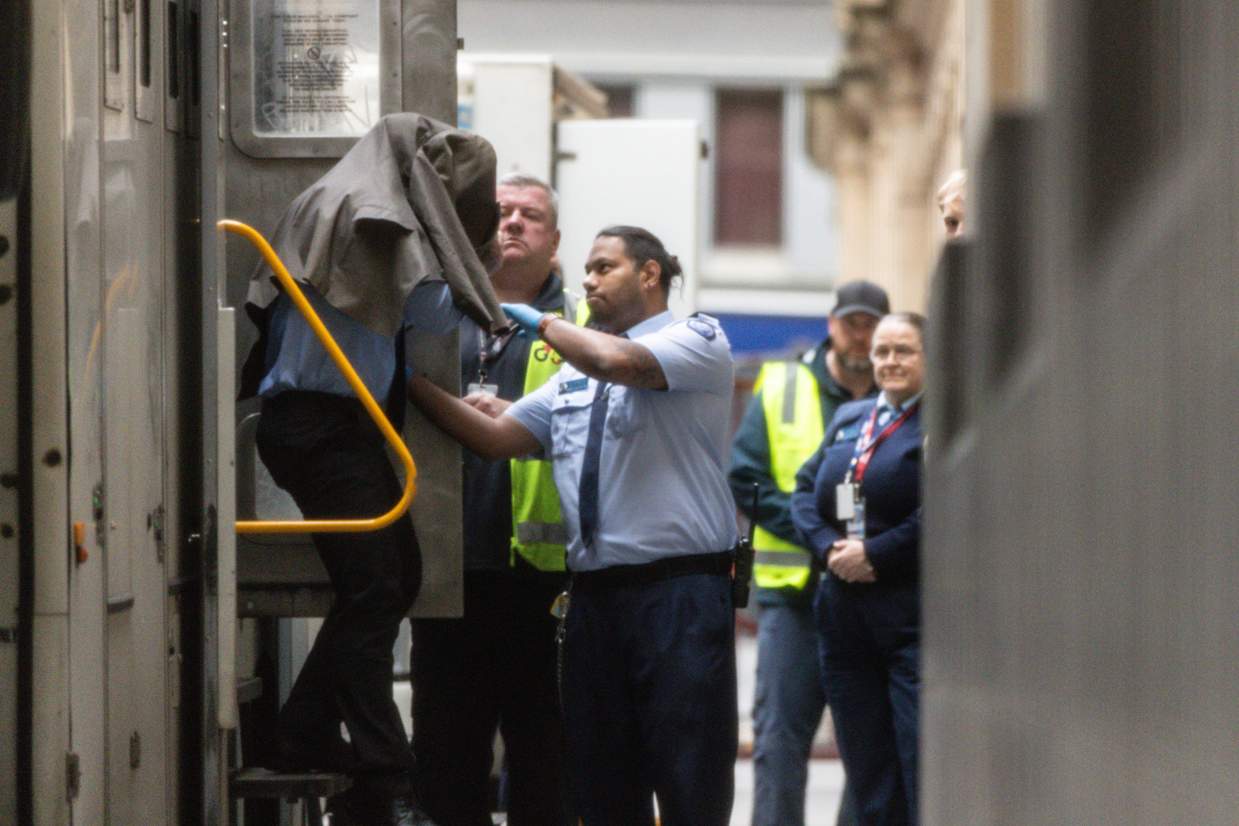 A person with their head covered by a jacket is escorted from a car by security.