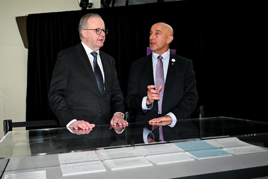 Two men in suits standing behind a display case with stacks of paper talking to each other
