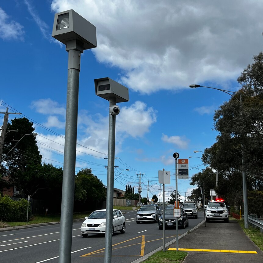 Two speed cameras pointed at a road, with a police car with its lights on parked in the near distance.