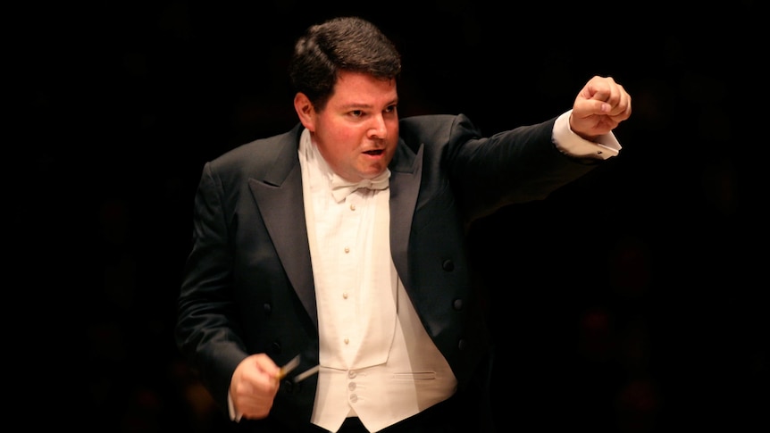 Andrew Litton in a tuxedo conducting with his left arm up in the air.