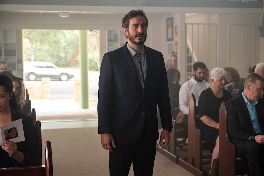 Ryan Corr as Charlie, standing in a church, people behind him in pews. He has a suit on.