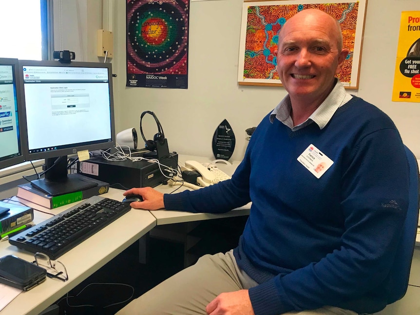 A man sits at a desk with a computer and Aboriginal artworks on the wall behind him.