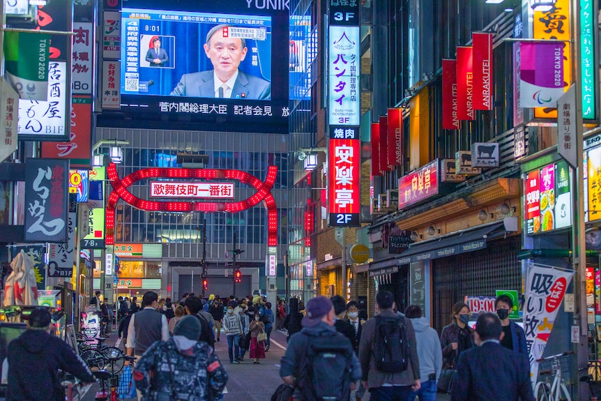 A street in Japan at night, with PM Yoshihide Suga speaking on a large TV above