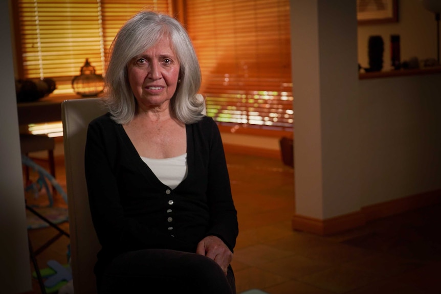 A woman sits facing the camera. She has grey hair and is wearing a black cardigan.