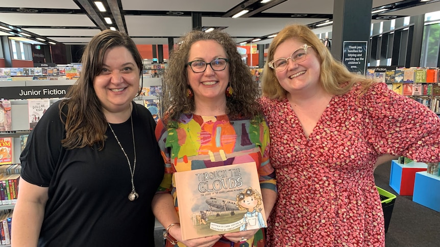 Three smiling women stand in a library, woman in middle, wears colourful dress, glasses, holds book.