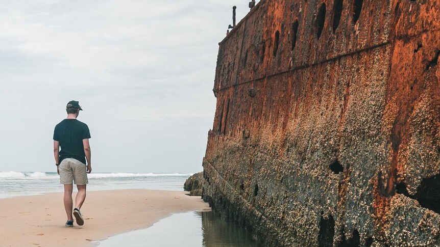 Man walking beside rusted shipwreck on beach