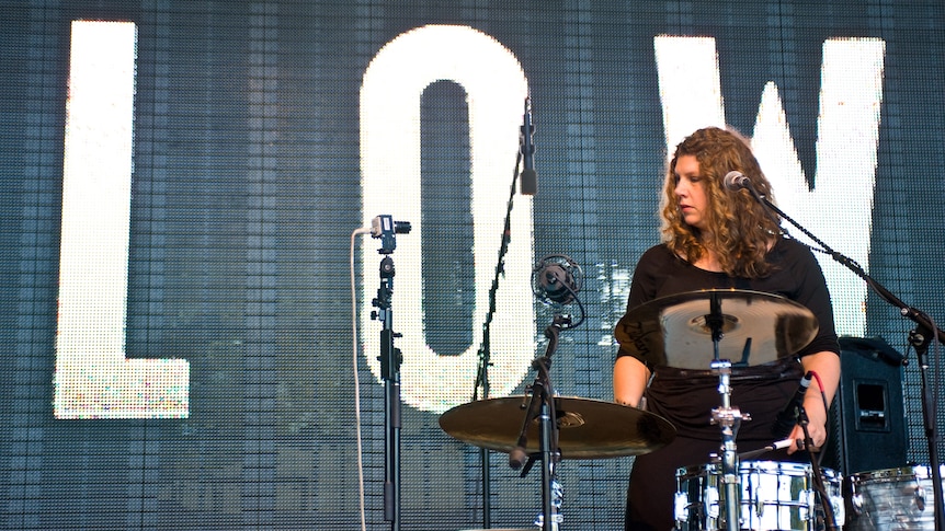 Mimi Parker stands at a drum set wearing black. Behind her the word LOW is written on a big screen