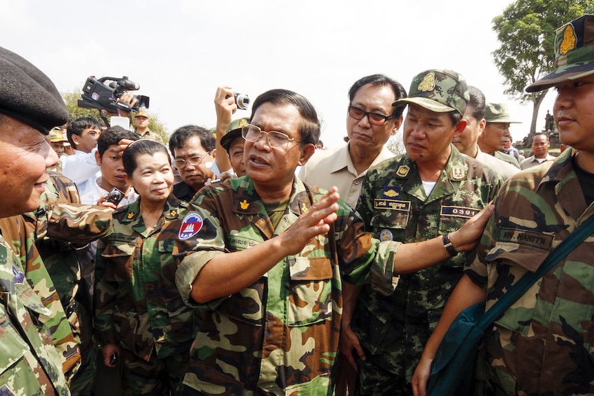 Hun Sen, wearing a military uniform, gestures while speaking to soldiers around him.