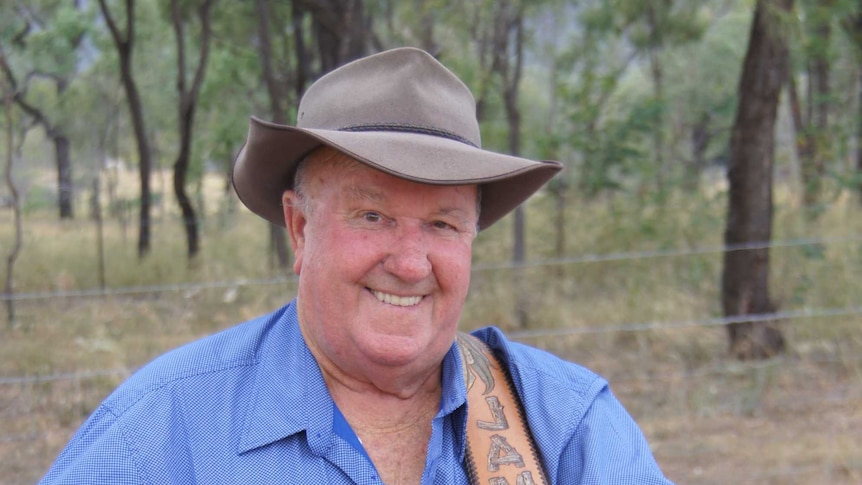 A smiling man in a blue shirt standing in front of trees and a barbed wire fence wearing a hat and holding his guitar.