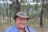 A smiling man in a blue shirt standing in front of trees and a barbed wire fence wearing a hat and holding his guitar.