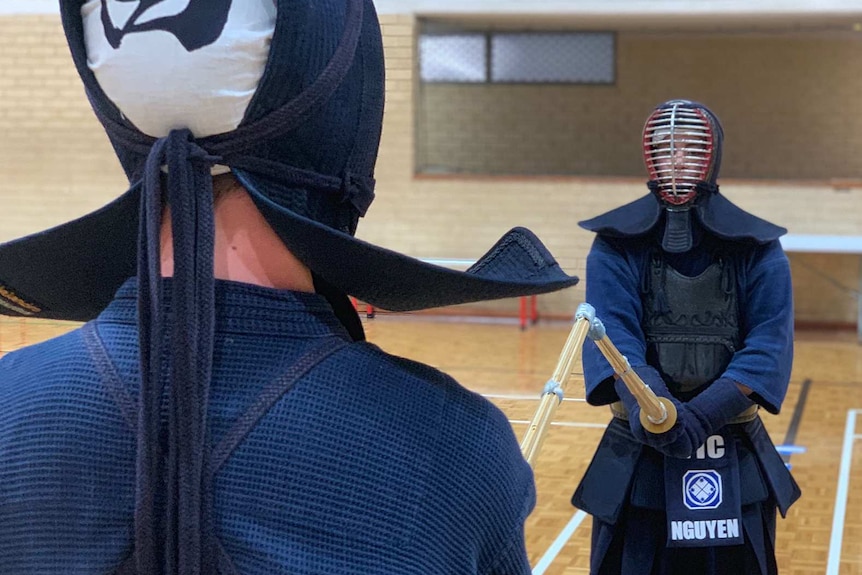 Two men in navy blue uniforms and headguards hold swords and face off in a gymnasium.