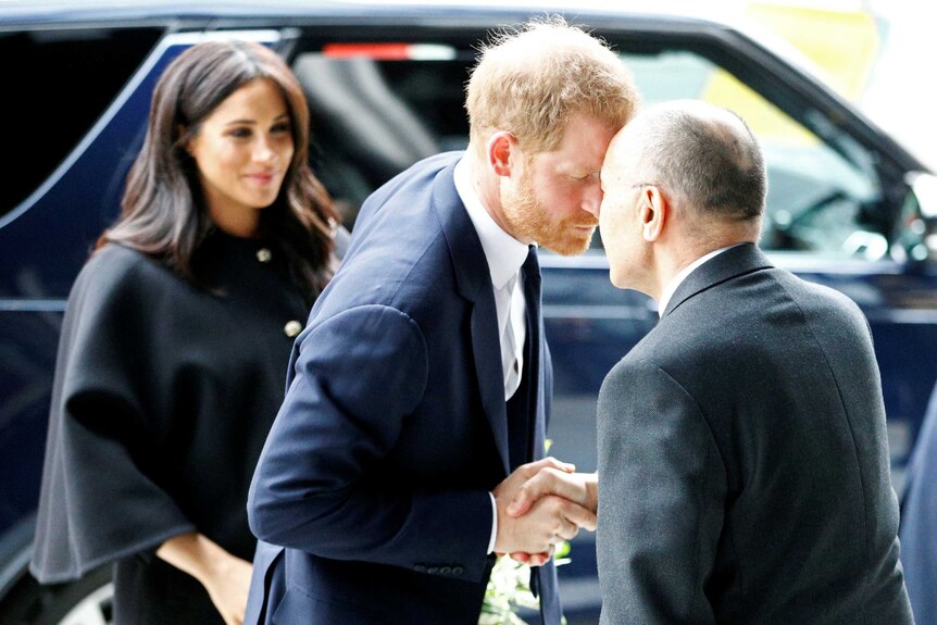 Prince Harry shakes hands and rubs noses with a man in a suit while Meghan Markle looks on in the background