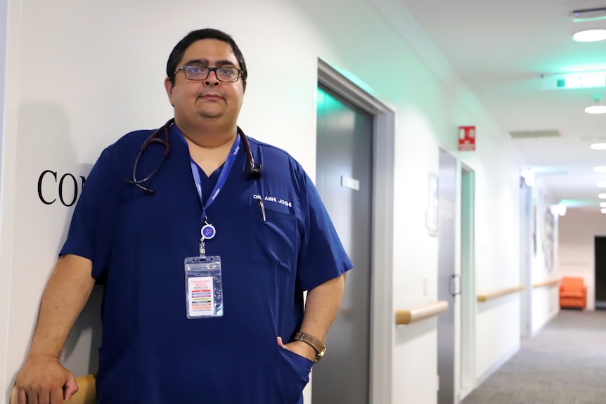doctor with glasses in navy blue scrubs stands in hospital corridor 