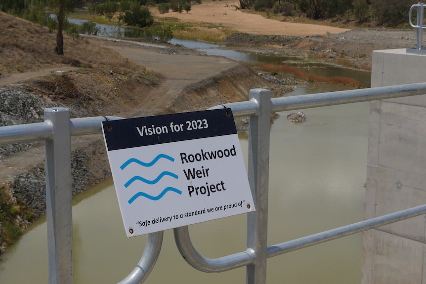 A metal fence in front of water with a sign that says "Vision for 2023, Rookwood Weir Project"