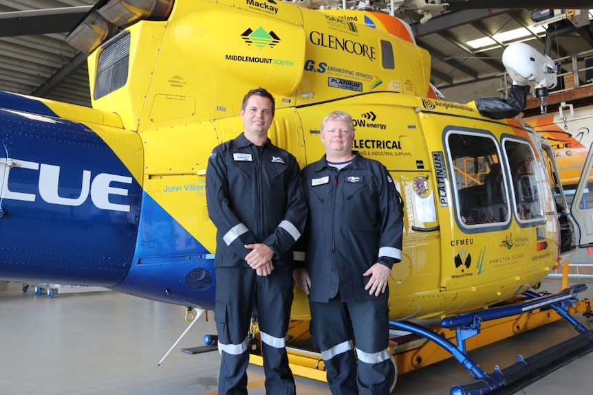 Ben McCauley and Kevin Berry stand in front of the RACQ rescue helicopter in its hanger.