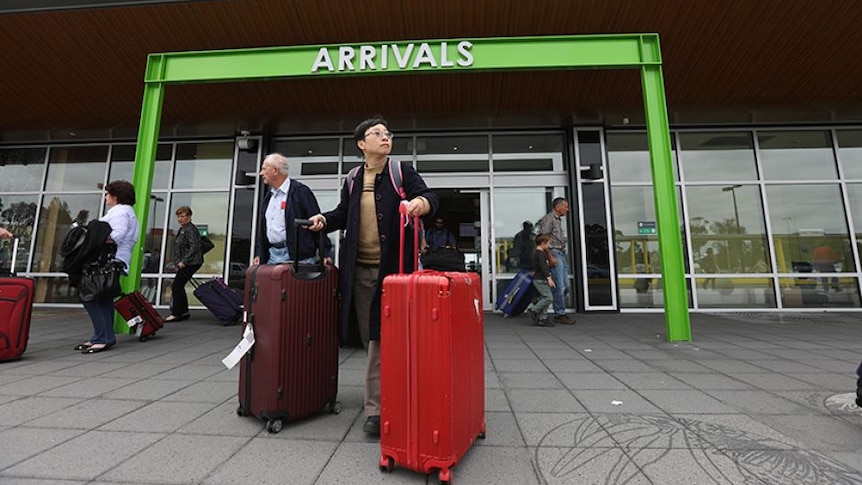 An Asian woman in black coat wheels two big suitcases outside the arrivals door of an airport