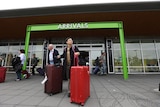 An Asian woman in black coat wheels two big suitcases outside the arrivals door of an airport