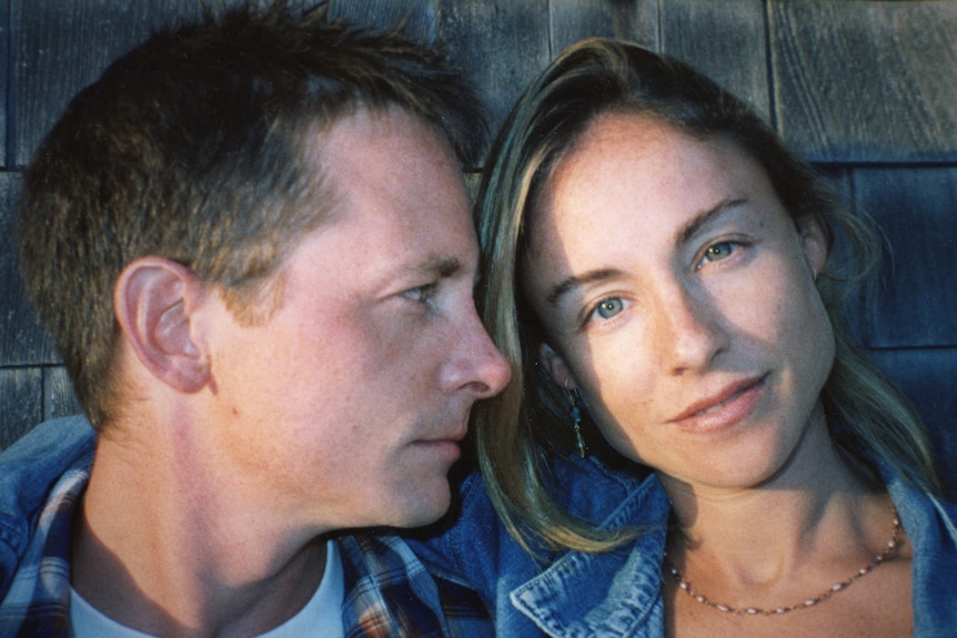 A young couple, both wearing denim jackets, lying together. The man is gazing across at the woman, who looks at the camera.