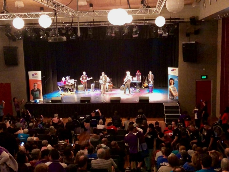 View of a stage from the rear of the theatre showing lots of people sitting while a band performs