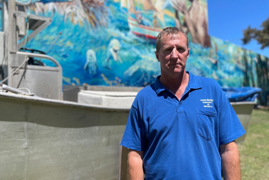 A man in a blue shirt leans against a small boat looking serious