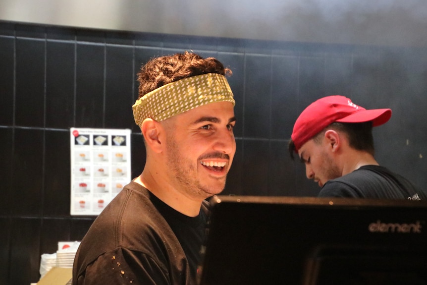 Man in green bandana standing behind a computer in a kitchen with cook working behind him.