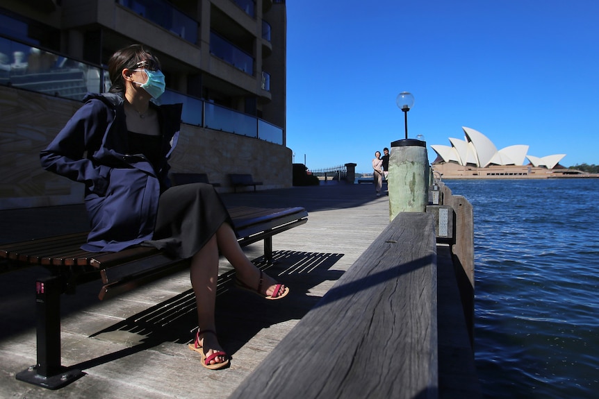 a woman wearing a mask sitting on a bench at a wharf opposite the opera house sydney harbpur