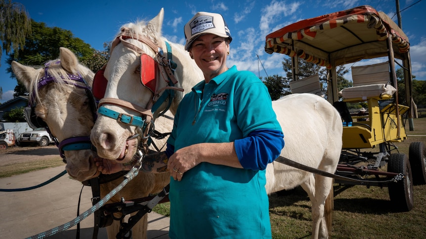 A woman in a blue top and white cap smiles next two her two white ponies harnessed to a yellow wagon
