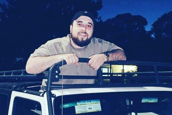 Michael Tsahrelias wears a cream t-shirt and black cap and stands on the back of a ute, smiling, at night.
