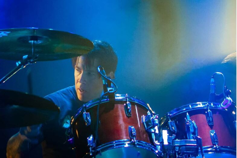 A blue-lit drummer plays while looking to the left of camera 